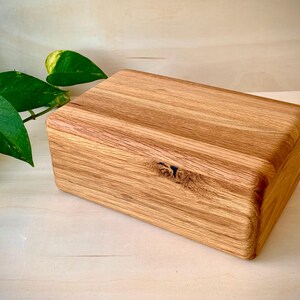 May include: A rectangular wooden box crafted from light brown oak with a visible natural wood grain pattern. The box features a smooth, rounded edge design and a prominent dark knot detail on the front panel, showcasing the organic texture of the wood.