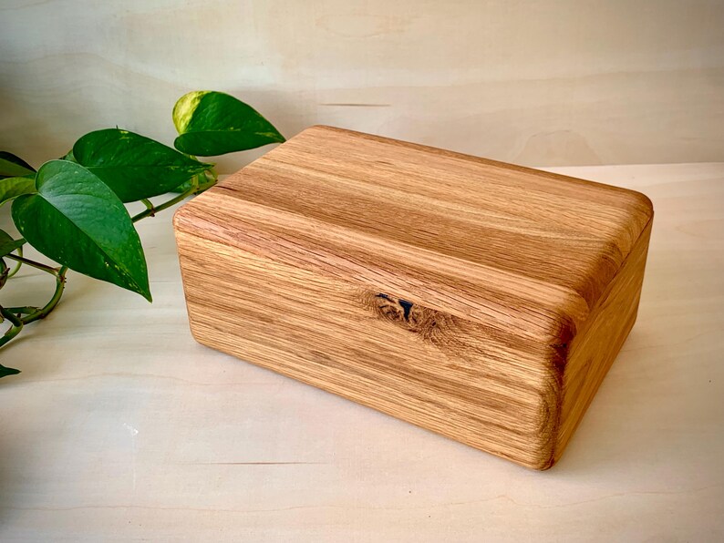 May include: A rectangular wooden box crafted from light brown oak with a visible natural wood grain pattern. The box features a smooth, rounded edge design and a prominent dark knot detail on the front panel, showcasing the organic texture of the wood.