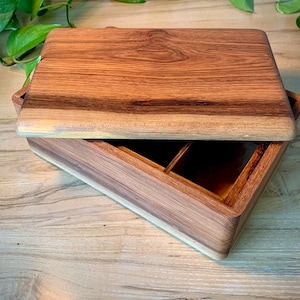 May include: A rectangular wooden box with a natural grain pattern and a removable lid. The box features a smooth, polished finish in shades of brown and tan. Inside, the container is divided into two separate compartments by a central divider.