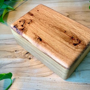 May include: A rectangular wooden box featuring a natural light brown finish with prominent wood grain patterns and dark knots on the top lid. The box has smooth, rounded edges and a polished surface, showcasing the organic texture of the wood.