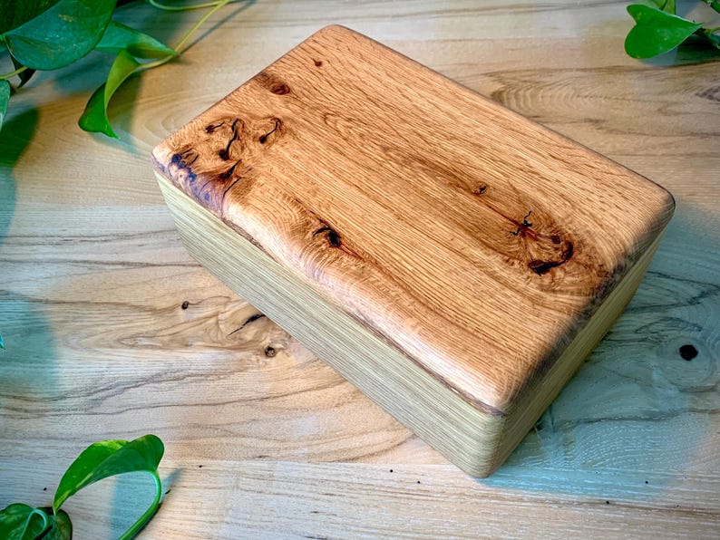 May include: A rectangular wooden box featuring a natural light brown finish with prominent wood grain patterns and dark knots on the top lid. The box has smooth, rounded edges and a polished surface, showcasing the organic texture of the wood.