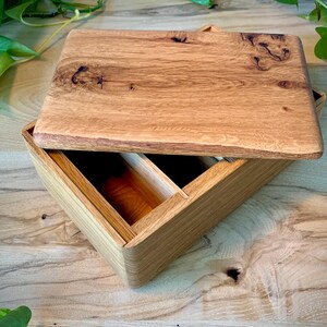 May include: A rectangular wooden box with a removable lid featuring a prominent natural wood grain pattern and dark knots. The interior of the box is divided into two sections, showcasing the light brown color and smooth texture of the wood.