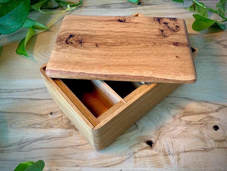 May include: A rectangular wooden box with a removable lid featuring a prominent natural wood grain pattern and dark knots. The interior of the box is divided into two sections, showcasing the light brown color and smooth texture of the wood.