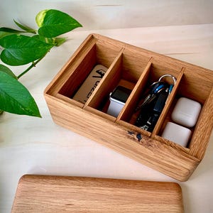 May include: A rectangular wooden organizer box with three internal compartments. The box contains a white rectangular card, a smartwatch with a white band, a set of metal keys on a ring, and two white square wireless earbud cases.