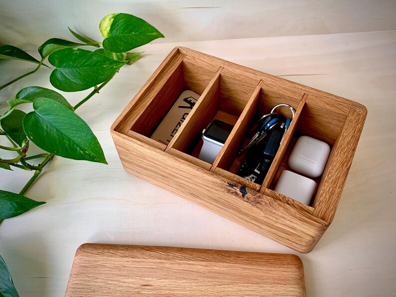 May include: A rectangular wooden organizer box with three internal compartments. The box contains a white rectangular card, a smartwatch with a white band, a set of metal keys on a ring, and two white square wireless earbud cases.