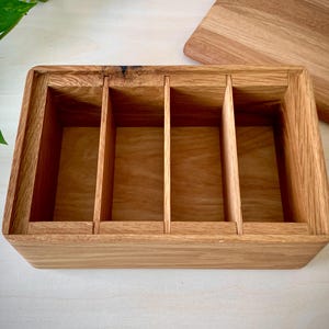 May include: A rectangular wooden storage box crafted from light brown oak, featuring four distinct interior compartments separated by three vertical dividers. The box is open at the top, revealing the natural wood grain and a smooth finish.