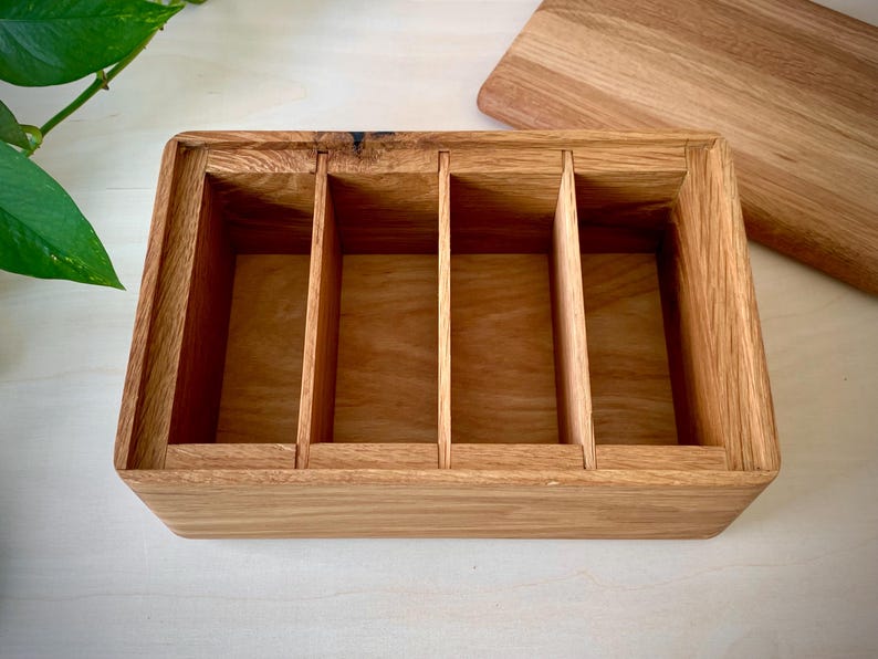 May include: A rectangular wooden storage box crafted from light brown oak, featuring four distinct interior compartments separated by three vertical dividers. The box is open at the top, revealing the natural wood grain and a smooth finish.