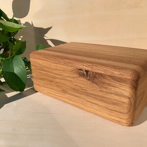 May include: A rectangular wooden box with a natural light brown grain pattern and a visible dark knot on the front panel. The box features rounded edges and a smooth finish, showcasing the organic texture and variations of the wood material.