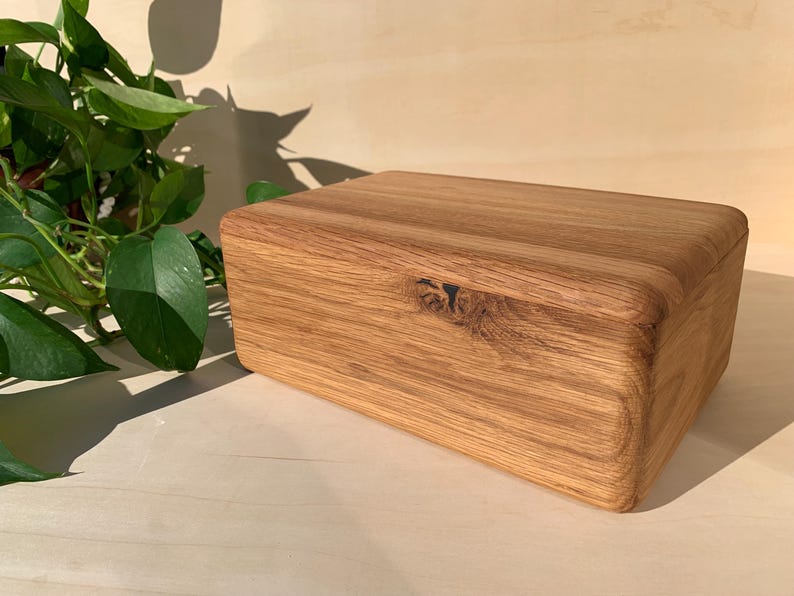 May include: A rectangular wooden box with a natural light brown grain pattern and a visible dark knot on the front panel. The box features rounded edges and a smooth finish, showcasing the organic texture and variations of the wood material.