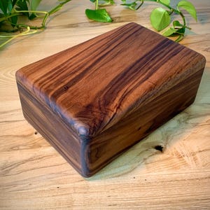 May include: A rectangular wooden box crafted from dark brown timber with prominent, swirling grain patterns. The box features a smooth, polished finish and a lid that sits flush with the base, showcasing the natural variations in the wood's color.