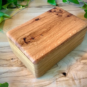 May include: A rectangular wooden box with a lid featuring a prominent natural wood grain pattern and a dark knot detail. The box has smooth, rounded edges and a light brown finish, showcasing the organic texture and variations of the wood.