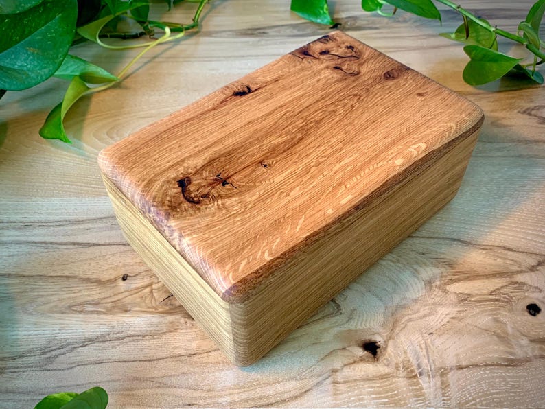 May include: A rectangular wooden box with a lid featuring a prominent natural wood grain pattern and a dark knot detail. The box has smooth, rounded edges and a light brown finish, showcasing the organic texture and variations of the wood.