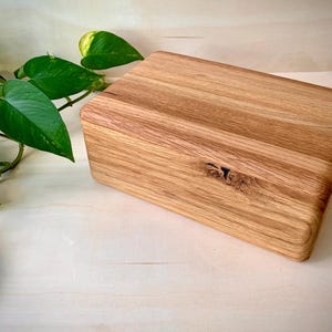 May include: A rectangular wooden box crafted from light-colored oak with a visible natural wood grain pattern. The box features a smooth, rounded top edge and a small, dark knot detail on the front face, showcasing the organic texture of the wood.