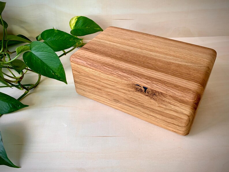 May include: A rectangular wooden box crafted from light-colored oak with a visible natural wood grain pattern. The box features a smooth, rounded top edge and a small, dark knot detail on the front face, showcasing the organic texture of the wood.