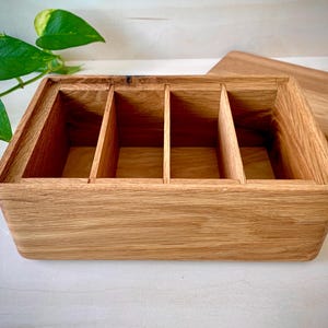 May include: A rectangular wooden storage box featuring a natural light brown wood grain finish. The interior is divided into four equal sections by three vertical wooden partitions. A separate matching wooden lid is positioned to the right side.