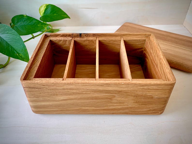 May include: A rectangular wooden storage box featuring a natural light brown wood grain finish. The interior is divided into four equal sections by three vertical wooden partitions. A separate matching wooden lid is positioned to the right side.