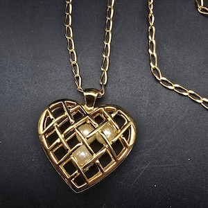 May include: Gold-toned necklace featuring a heart-shaped pendant with a lattice design. The pendant holds three faux pearls. The chain is a classic link style. The jewelry is displayed against a dark background.