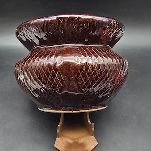 May include: A brown glazed ceramic pot with a diamond pattern and a wide, flared rim. The pot is supported by a wooden stand. The pot's surface has a glossy finish, reflecting light. The overall shape is rounded and bulbous.