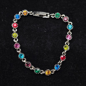 May include: A silver-toned metal chain bracelet featuring a series of round, multi-colored faceted gemstones in shades of red, blue, green, yellow, and pink. The bracelet is secured with a rectangular metal clasp mechanism at the top center.