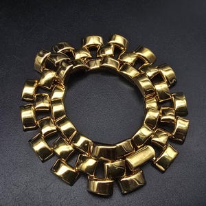 May include: A circular gold-tone bracelet featuring a multi-layered, interlocking link design. The piece is composed of polished, curved rectangular metal segments arranged in a dense, concentric pattern that creates a textured, reflective surface.