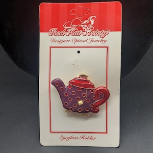 May include: A Red Hat Society Designer Optical Jewelry eyeglass holder pin shaped like a teapot. The teapot is purple with red polka dots, a red lid, and a red handle. It is attached to a white and red display card with the text Red Hat Society.