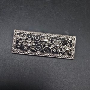 May include: A rectangular silver-toned brooch with an intricate floral and swirl design. The brooch features a series of stylized flowers with circular accents and is set against a dark background. The edges are lined with small, round embellishments.