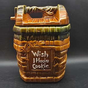 May include: A ceramic cookie jar shaped like a well, with a handle on top and a lid. The jar is primarily brown with green and tan accents. The words "Wish I Hada Cookie" are displayed on the front in a white rectangular frame.