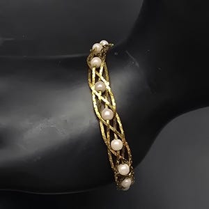 May include: A gold-tone bracelet featuring a textured, crisscross lattice design. The metal structure is accented with small, round, white pearls set at regular intervals along the center of the woven pattern, creating a continuous decorative band.
