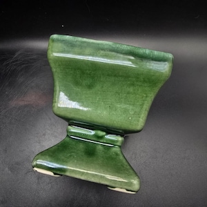 May include: A square-shaped ceramic planter with a glossy, deep forest green glaze. The vessel features a flared top rim, a tapered midsection, and a wide, stable pedestal base. The surface shows subtle variations in color intensity and texture.