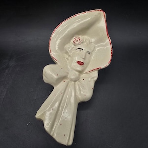 May include: A vintage ceramic wall hanging in the shape of a woman wearing a wide-brimmed hat. The figure is painted in off-white with red accents on the hat brim, lips, and small details. A rose adorns the hat.