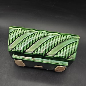 May include: A rectangular, green ceramic planter with a glossy finish. The planter features diagonal stripes in varying shades of green, creating a textured appearance. The base has small, light-colored feet. A decorative item for plants.