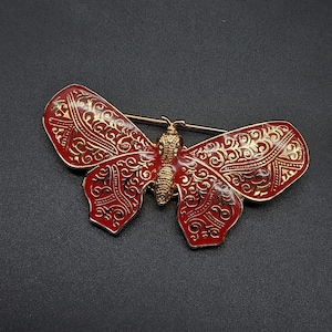 May include: A butterfly-shaped brooch featuring a vibrant red enamel finish with intricate gold-toned filigree scrollwork patterns across the wings. The central body is textured with a gold-toned finish, and a horizontal pin mechanism is visible.
