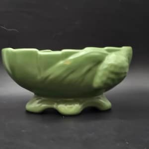 May include: A green ceramic planter with a scalloped rim and a textured, leaf-like design. The planter sits on three curved feet. The overall shape is a shallow bowl, suitable for small plants or decorative use.
