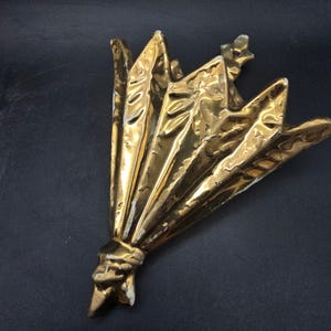 May include: A gold-colored, fan-shaped decorative wall hanging. The piece has a textured surface and a tied base, resembling a bouquet. The design features multiple pointed sections, creating a layered effect. The overall aesthetic is vintage.