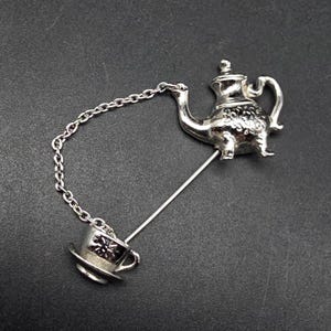May include: A silver-toned brooch featuring a teapot and teacup design. The teapot has intricate detailing and a handle, connected by a chain to a teacup on a saucer. The brooch is attached with a long pin, suitable for pinning on clothing or accessories.
