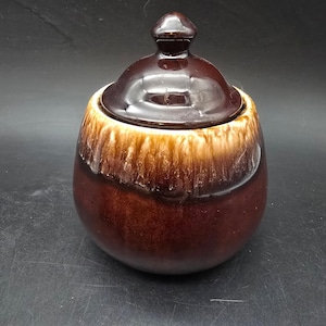 May include: A brown ceramic sugar bowl with a lid. The bowl has a rounded shape with a two-tone glaze, transitioning from a light tan to a dark brown. The lid is a matching dark brown with a small knob on top.