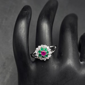 May include: A silver ring featuring a floral design with a central red gemstone surrounded by green and white stones. The ring is set on a black surface, showcasing its intricate details and vibrant colors.