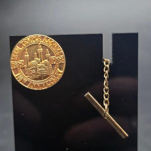 May include: A gold-colored pin with the text "TEMPLE AAONMS 1895 1995 100TH ANNIVERSARY" encircling a detailed building illustration. The pin is attached to a black display with a gold chain and bar.