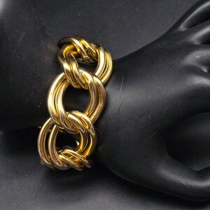 May include: A gold-toned chain link bracelet. The bracelet features a series of large, interlocking oval links. The links have a twisted rope-like texture. The bracelet is displayed on a black surface.