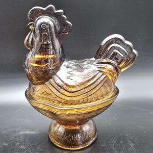 May include: Amber-colored glass rooster figurine with intricate detailing. The rooster is perched on a pedestal base, and features a detailed comb, wattle, and tail feathers. The glass has a textured appearance, and the design is likely vintage.