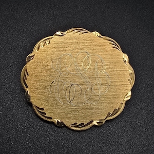 May include: A gold-colored, oval-shaped vintage compact with an engraved monogram. The compact has a decorative, scalloped edge with leaf-like accents. The surface has a brushed texture, and the monogram appears to be the letters "E" and "S".