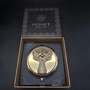 May include: A gold-toned compact mirror with an angel design, presented in a black box. The angel features detailed wings and a halo. The box is marked with the brand name "MONET" and the text "SINCE 1929".
