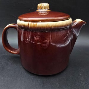 May include: A brown ceramic teapot with a lid and handle. The teapot has a glossy finish with a lighter tan band around the rim. The lid has a small, decorative knob. The spout is on the right side of the teapot.
