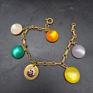 May include: Vintage gold-tone charm bracelet featuring a series of colorful, round charms. The charms include a green, orange, yellow, and lavender cabochon, plus a round charm with a United Air Corps emblem.