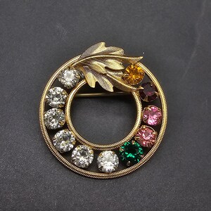 May include: A gold-toned brooch with a circular design featuring a leaf and colorful rhinestones. The rhinestones are in shades of pink, green, red, and orange.