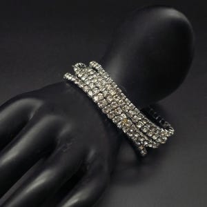 May include: A multi-row bracelet featuring clear, faceted rhinestones set in a silver-toned metal frame. The bracelet is designed as a flexible wrap style, consisting of several parallel strands of sparkling stones that encircle the wrist area.