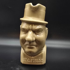May include: A tan ceramic pitcher in the shape of W.C. Fields' head, complete with a top hat. The pitcher has a stern expression and the name "W.C. FIELDS" is printed on the base. The pitcher is a collectible item.