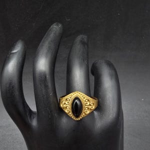 May include: A gold-colored ring with an oval, black stone set in the center. The ring has an ornate design with swirling patterns around the stone. The ring is displayed on a black hand.