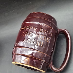 May include: A brown ceramic beer mug with a large handle. The mug has a cylindrical shape with horizontal ridges and the words "HAPPY DAYS ARE HERE AGAIN" embossed on the front. The mug is made of a glossy, dark brown ceramic.