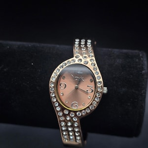 May include: A gold-toned wristwatch with a blush-colored face and a band adorned with clear crystals. The watch face features the word "York" and the numbers 3, 6, and 9. The watch is displayed on a black surface.